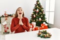 Caucasian young blonde woman sitting on the table by christmas tree begging and praying with hands together with hope expression Royalty Free Stock Photo