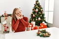 Caucasian young blonde woman sitting on the table by christmas tree sleeping tired dreaming and posing with hands together while