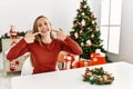 Caucasian young blonde woman sitting on the table by christmas tree smiling cheerful showing and pointing with fingers teeth and