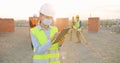 Caucasian young good-looking woman engineer in helmet and medical mask standing at constructing side, using tablet