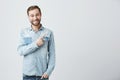 Caucasian young male in denim shirt grins at camera, indicates at copy space, advertises something. Happy bearded man