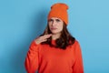 Caucasian young woman with disgusted expression feels disgust keeping hand near frowning face wearing casual orange jumper and