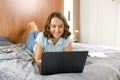 Caucasian young woman lying down on the cozy bed in cozy bright bedroom and using laptop computer, chatting online Royalty Free Stock Photo