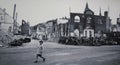 An antique photo shows young child in school uniform crossing road with destroyed building and cars in his back. Copy of film