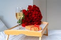 Celebrating Saint Valentine`s Day with bouquet of red roses and glass of champagne on tray on bed. Royalty Free Stock Photo