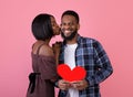 Celebrating Valentine`s Day. Lovely black girl in dress kissing her happy boyfriend with red heart over pink background Royalty Free Stock Photo