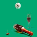 Celebration arrangement of disco ball decorations above red berries, bottle of champagne and crystal glass. Vivid green background