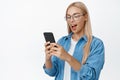 Cellular and smartphone technology. Young cute girl in glasses looking at her mobile phone with surprised face, standing Royalty Free Stock Photo
