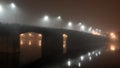 Cement bridge in thick fog. Street lights illuminate road at night, reflecting in calm dark river water. Mystical atmosphere. Royalty Free Stock Photo