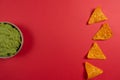 Ceramic bowl with guacamole on a red background with copy space in the center and some corn triangles on the side