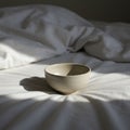 A ceramic bowl rests on a white, rumpled bed with sunlight casting soft shadows. Th Royalty Free Stock Photo