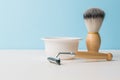 A ceramic bowl, a shaving brush and a razor with a wooden handle on a white table. Space for the text