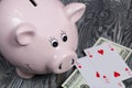 Ceramic piggy bank in the form of a pink pig. Nearby are dollar bills and playing cards. Against the background of aged wooden Royalty Free Stock Photo