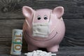 Ceramic piggy bank in pink. With a gauze bandage. In front of her is a handful of white pills. Lay dollar bills, rolled up into a Royalty Free Stock Photo