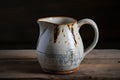 A ceramic pitcher with a handle and a spout, white with a blue speckled pattern and a brown rim, on a wooden surface with a dark