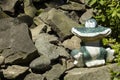 Ceramic smiling frog sitting on a pile of stones and next to green plants in a garden