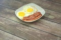 A ceramic white plate with two fried eggs and two slices of fried bacon stands on a wooden table. Close-up Royalty Free Stock Photo