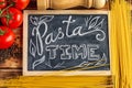 Chalk board sign Pasta Time on a wooden tabletop with raw spaghetti, fresh tomatoes and black pepper