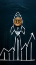 Chalk rocket with Bitcoin coin on blackboard, ascending with background line graph