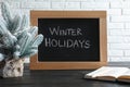 Chalkboard with phrase Winter Holidays, small Christmas tree and book on black wooden table near white brick wall Royalty Free Stock Photo