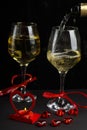 Champagne is poured into glasses on a black background next to a heart-shaped candlestick with a burning candle and a box with a