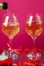 Champagne is poured into glasses on a pink background next to a heart candlestick with a burning candle and a box with a
