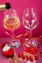 Champagne is poured into glasses on a pink background next to a heart candlestick with a burning candle and stones, hearts and a