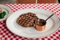 Charcoal grilled wagyu Ribeye steak served with BBQ sauce and baked potato in white plate on red and white pattern tablecloth