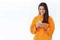 Charismatic pretty brunette girl in orange hoodie smiling and looking mobile phone screen as texting, messaging with