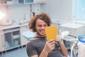 Charismatic smiling young man on the dentist chair , after a oral hygiene procedure take a mirror and looking at his Royalty Free Stock Photo