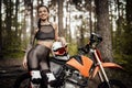 Charismatic tattooed young racer girl smiling and looking on the camera while leaning on her bike in the woods