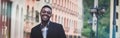 Charming african young businessman in suit posing next to office building, smiling africa-american entrepreneur having break after Royalty Free Stock Photo