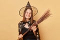 Charming attractive young woman wizard wearing witch costume holding in hand broom  over beige background expressing Royalty Free Stock Photo