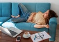 Charming beautiful young girl in glasses with curly hair fell asleep on a blue sofa at home in front of a laptop, notepad and a Royalty Free Stock Photo