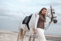 Charming and beautiful young woman wearing stylish jockey outfit is holding the reins and posing with the white horse