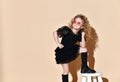 Blonde curly kid in orange sunglasses, necklace, black dress and boots. She put her foot on tabouret, posing on beige background