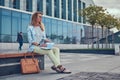 Charming blonde female in modern clothes, studying with a book, sitting on a bench in the park against a skyscraper. Royalty Free Stock Photo