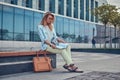 Charming blonde female in modern clothes, studying with a book, sitting on a bench in the park against a skyscraper. Royalty Free Stock Photo