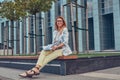 Charming blonde female in modern clothes, studying with a book, sitting on a bench in the park against a skyscraper. Royalty Free Stock Photo