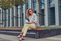 Charming blonde female in modern clothes, studying with a book, sitting on a bench in the park against a skyscraper. Royalty Free Stock Photo