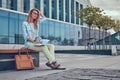 Charming blonde female in modern clothes, studying with a book, sitting on a bench in the park against a skyscraper. Royalty Free Stock Photo