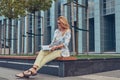 Charming blonde female in modern clothes, studying with a book, sitting on a bench in the park against a skyscraper. Royalty Free Stock Photo