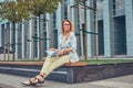 Charming blonde female in modern clothes, studying with a book, sitting on a bench in the park against a skyscraper. Royalty Free Stock Photo