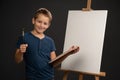 Charming boy in blue t-shirt looking at camera holding a palette with paints on the background of an easel with canvas