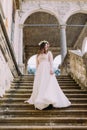 Charming bride in long white wedding dress and floral wreath going down by antique palace stone stairs Royalty Free Stock Photo