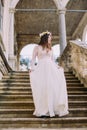 Charming bride in long white wedding dress and floral wreath going down by antique stone stairs Royalty Free Stock Photo