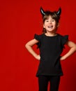 Little girl with devil horns, in black blouse and leggings. She laughing, hands on hips, posing on red background. Close up Royalty Free Stock Photo