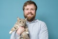 Charming Caucasian bearded man hugs his adorable cat, smiles and looks at camera, on a blue background. Concept of love