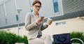 Charming caucasian businesswoman in glasses scrolling and taping on the smartphone while drinking coffee on the bench