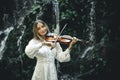 Charming Caucasian woman playing violin with closed eyes near waterfall. Music, art concept. Female with blond hair wearing white Royalty Free Stock Photo